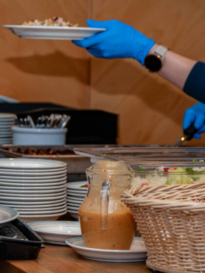 Buffet-pöytä, jossa on salaattia, leipää ja kastiketta. Taustalla henkilö tarjoilee ruokaa lautaselle sinisissä käsineissä. Kuvaaja Katja Rantavaara