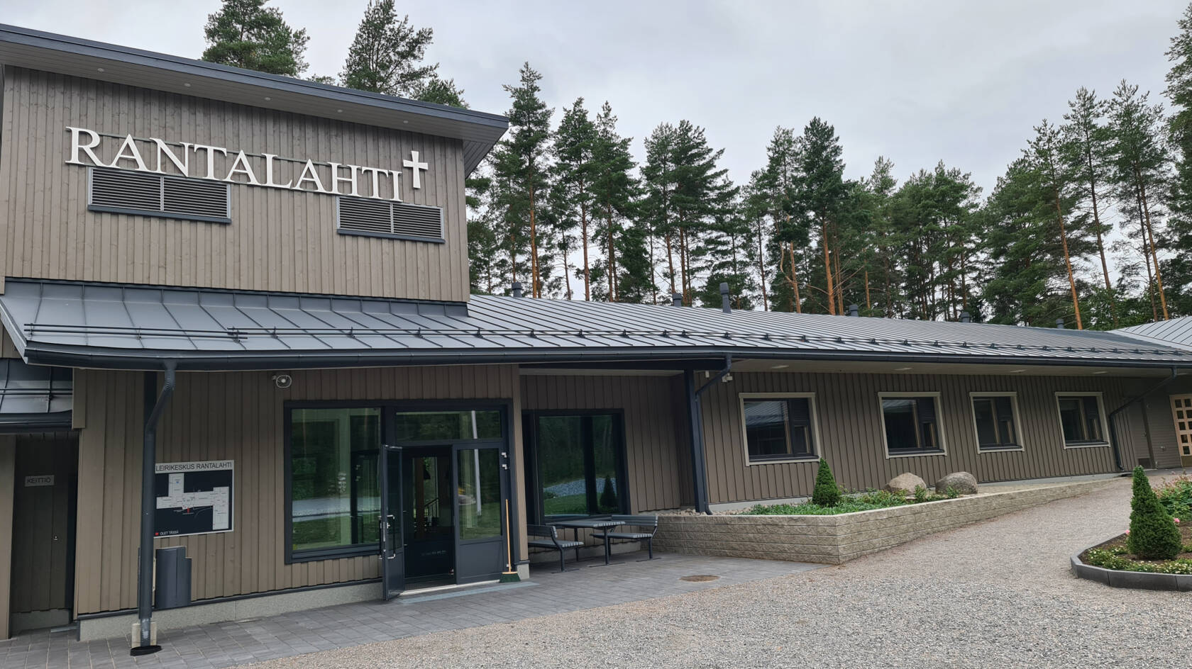 Harmaa leirikeskusrakennus, sisäänkäynti ja ikkunoita. Katon rajassa RANTALAHTI-teksti.