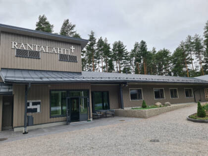 Harmaa leirikeskusrakennus, sisäänkäynti ja ikkunoita. Katon rajassa RANTALAHTI-teksti.