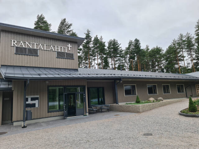 Harmaa leirikeskusrakennus, sisäänkäynti ja ikkunoita. Katon rajassa RANTALAHTI-teksti.