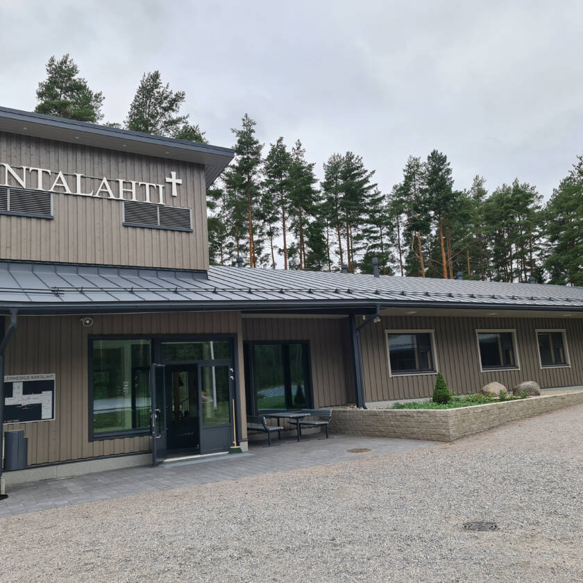 Harmaa leirikeskusrakennus, sisäänkäynti ja ikkunoita. Katon rajassa RANTALAHTI-teksti.