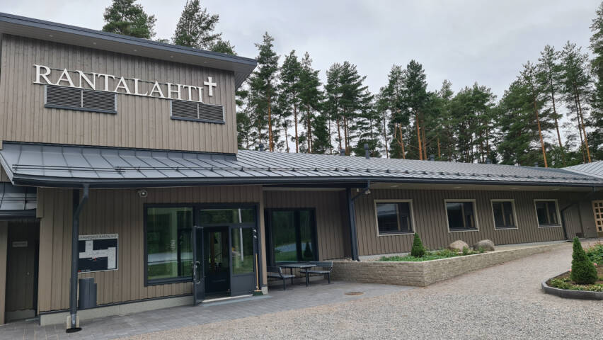 Harmaa leirikeskusrakennus, sisäänkäynti ja ikkunoita. Katon rajassa RANTALAHTI-teksti.