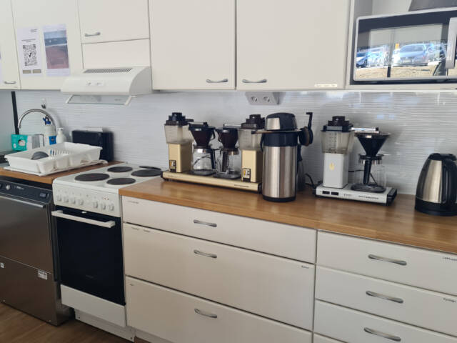 Kahvilan keittiötila, jossa työtasolla on useita kahvinkeittimiä ja termoskannuja. Alapuolella kaappeja ja liesi.