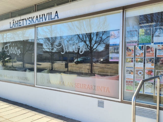 Kahvilan näyteikkuna ulkoa päin. Tekstit ”Lähetyskahvila Tavo” ja ”Janakkalan seurakunta” näkyvät ikkunassa.
